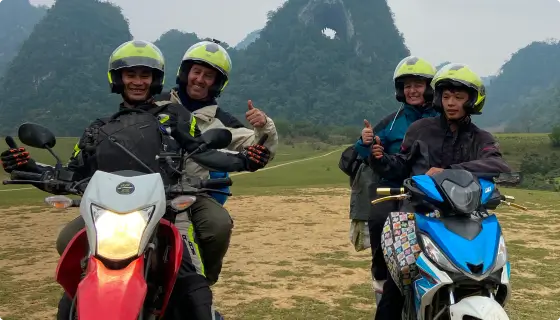 A smiling man and woman sitting on the back of motorcycles driven by tour guides, with mountains in the background