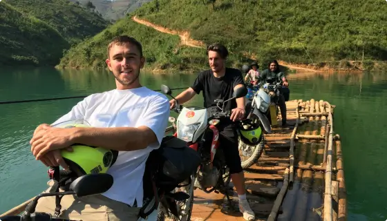 Four smiling young men sitting on rented Honda XR 150 motorcycles crossing a river in Vietnam on a wooden raft