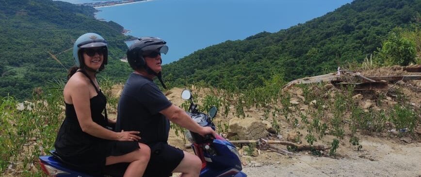 ALT TEXT; A smiling man and woman sitting on the back of motorcycles driven by tour guides, with mountains in the background